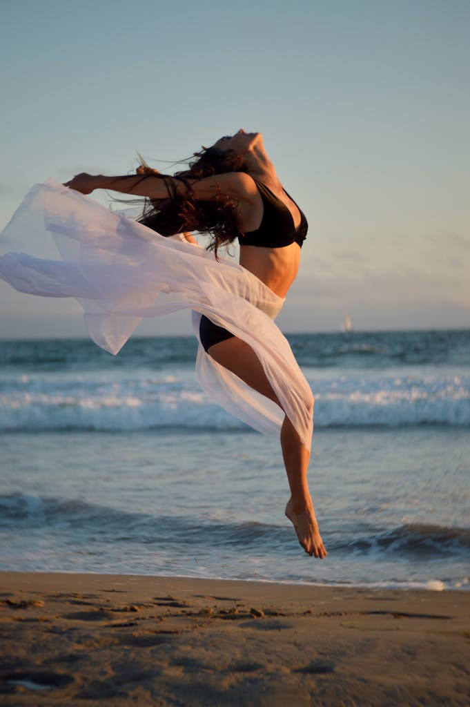 A carefree woman joyfully dances on the beach at sunset, showcasing freedom and elegance with the ocean backdrop.