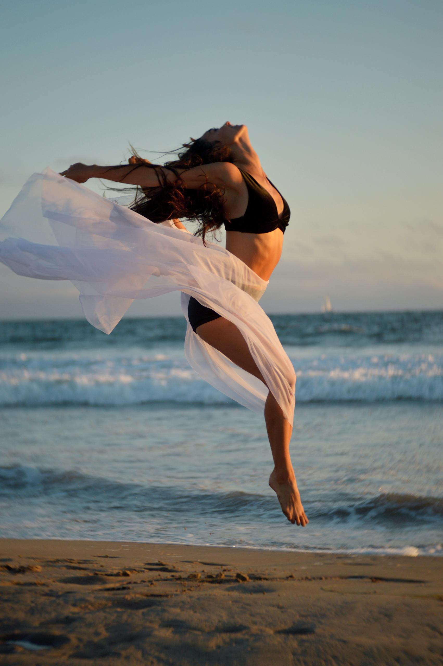 A carefree woman joyfully dances on the beach at sunset, showcasing freedom and elegance with the ocean backdrop.